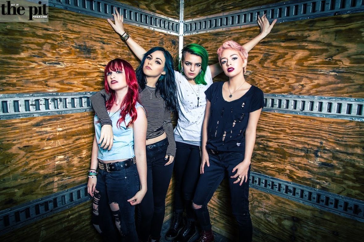 Four young women with colorful hairstyles posing confidently in a room with wooden panel walls.