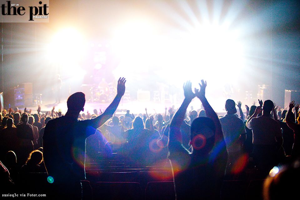 Audience members raising hands at a brightly lit concert, seen from behind with intense stage lights in the background.