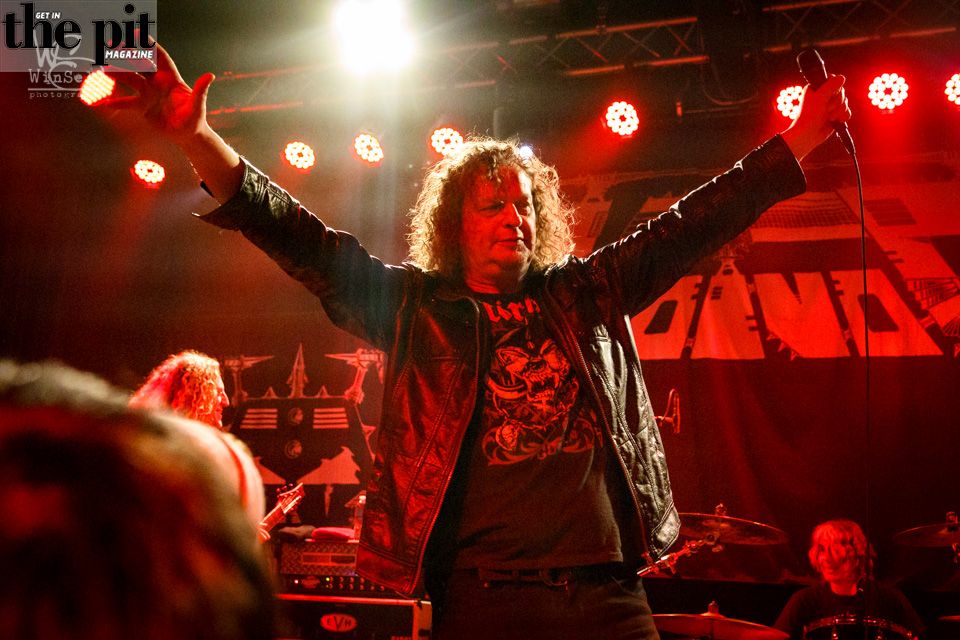 A man with curly hair raises his arms on stage at a rock concert, with red stage lights and band banners in the background.
