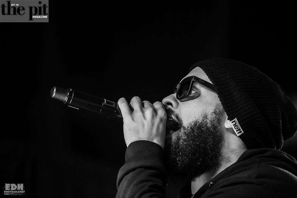 Black and white photo of a male singer in sunglasses and beanie performing into a microphone, with a logo of "the pit: magazine" in the background.