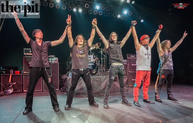 Five men from Ratt on stage at a concert, posing with their arms raised and holding hands up, with musical equipment in the background.