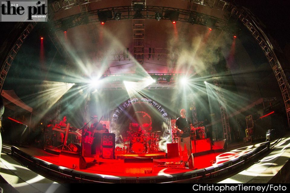Wide-angle photo of a Slightly Stoopid concert stage with vibrant green and white lighting, showcasing musicians performing with a drum set visible in the center.