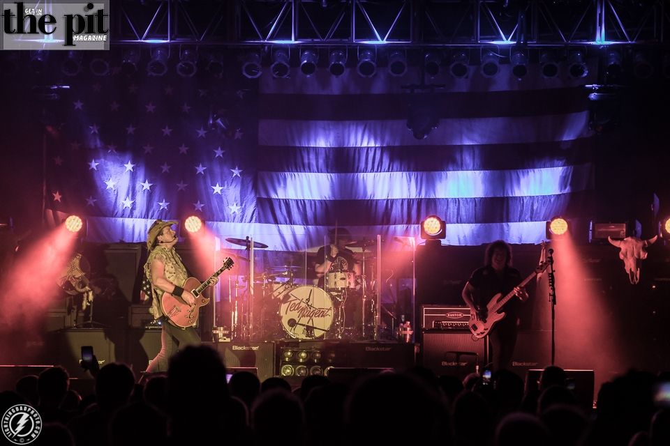 A rock band performs on stage, with a drummer at the back and two guitarists in the foreground, under lighting that highlights a Ted Nugent-themed American flag backdrop.