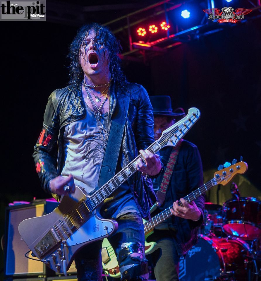 Rock musician Tom Keifer with black hair playing guitar on stage, wearing a leather jacket, under colorful stage lights. Another guitarist in the background.