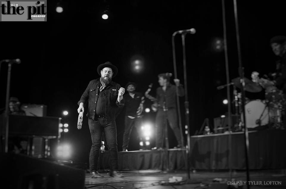 A musician in a hat playing guitar on stage with Nathaniel Rateliff and the Night Sweats in the background, in a monochrome live concert setting.