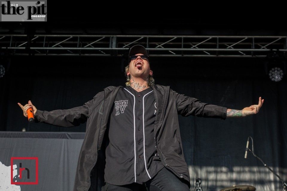 A male singer with tattoos, wearing sunglasses and a jacket, performs energetically at Dirt Fest with microphones in the background.