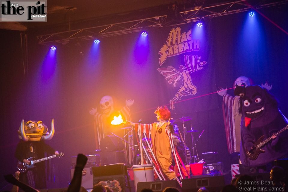Band members in costumes perform on stage, one with a flaming prop, under purple lighting with a "Mac Sabbath" banner in the background.