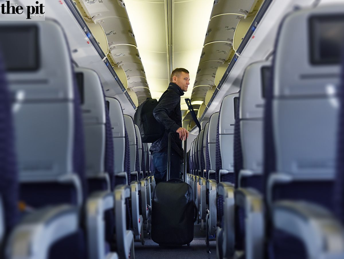 A passenger standing in an airplane aisle with an Incase x Kaskade backpack and rolling suitcase, looking towards the rear of the cabin.