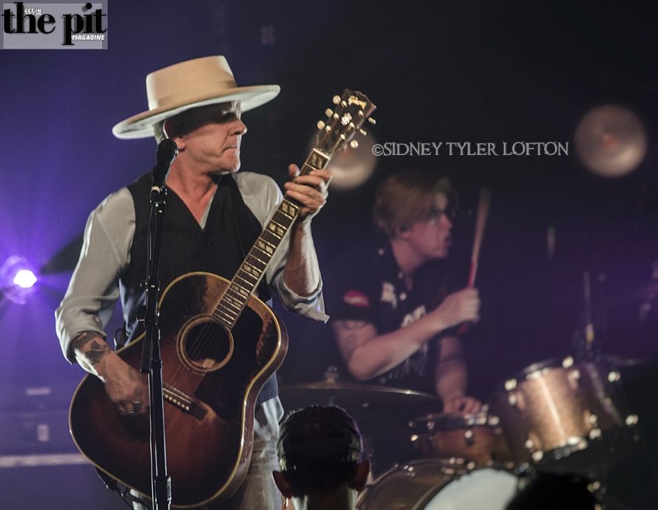 A male musician resembling Kiefer Sutherland in a white hat playing an acoustic guitar on stage, with a drummer blurred in the background. The image includes a watermark and magazine logo.