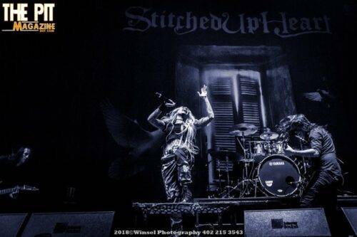 The Pit Magazine Pitcast! A band performs on stage; the vocalist gestures dramatically to the crowd, lit by moody blue stage lights, with a drummer and guitarist also visible. They are members of Stitched Up Heart.