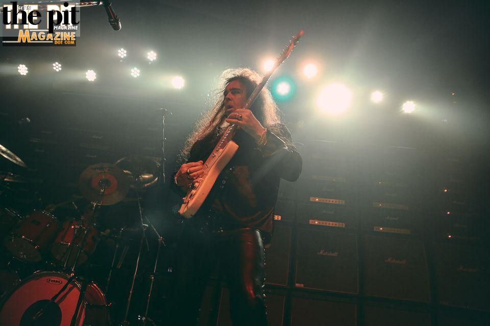 A musician with long curly hair resembling Yngwie Malmsteen playing an electric guitar on stage under blue and yellow stage lights, with drums and amplifiers in the background.