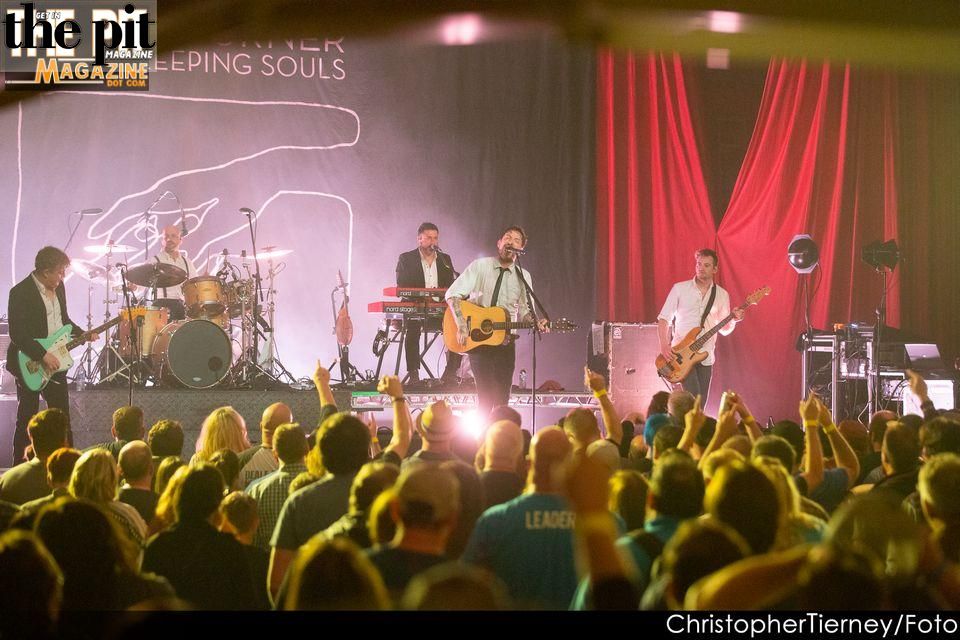 Frank Turner performing on stage in front of an energetic audience with logo and text overlay in the top left corner.