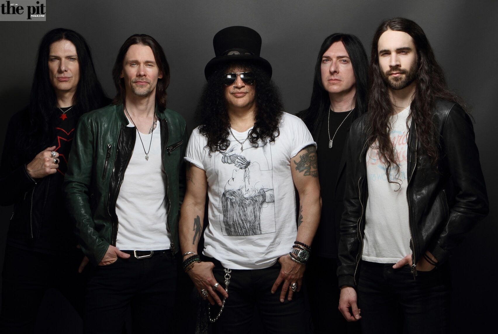 Five men from a rock band pose for a photo, wearing leather jackets and dark clothes, with Slash sporting a top hat.