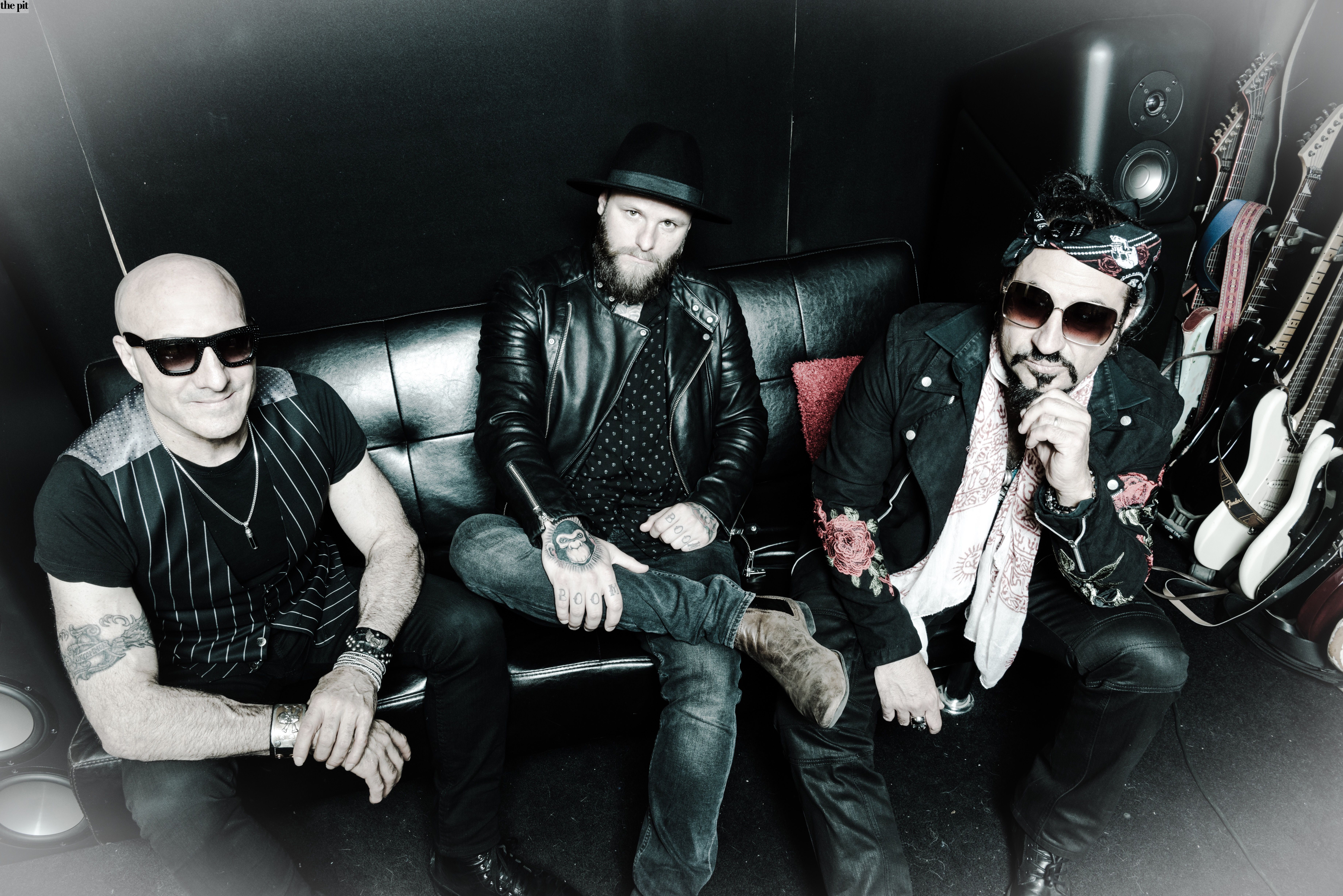 Three male musicians from Supersonic Blues Machine sitting on a black leather sofa in a recording studio, wearing edgy rock-style clothes and accessories.