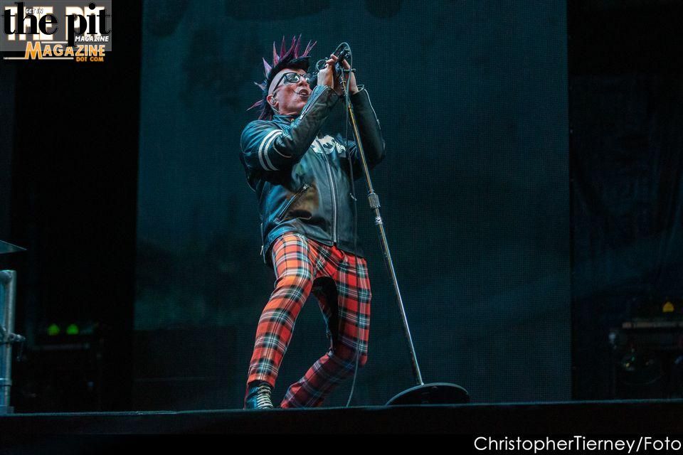 A male rock singer on stage, wearing red plaid pants and a gray jacket, passionately singing into a microphone with dramatic lighting, resembling the intense style of Tool.