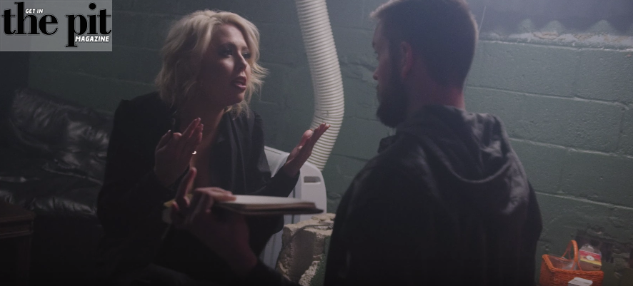 Two people having an intense discussion backstage, one holding a notebook. The scene is dimly lit in an industrial setting with visible ventilation ducts and Chloe Lowery participating in the conversation.
