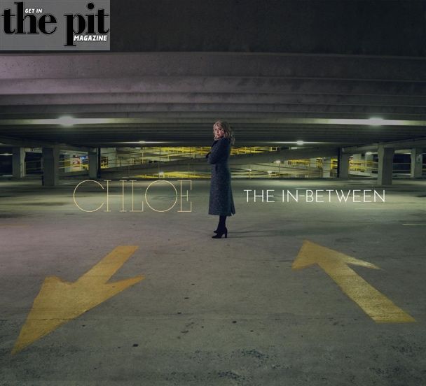 Woman in a coat standing in a parking garage with yellow directional arrows on the ground, under the title "Chloe Lowery the In-Between.