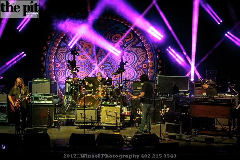 Gov't Mule performing on stage with purple stage lights and a large, colorful mandala backdrop.