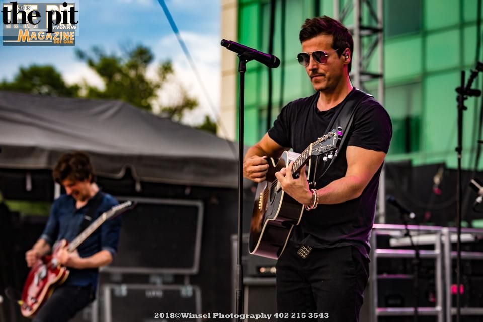 A male guitarist in sunglasses and a dark shirt strums an acoustic guitar onstage, while another musician from Our Lady Peace plays electric guitar in the background.