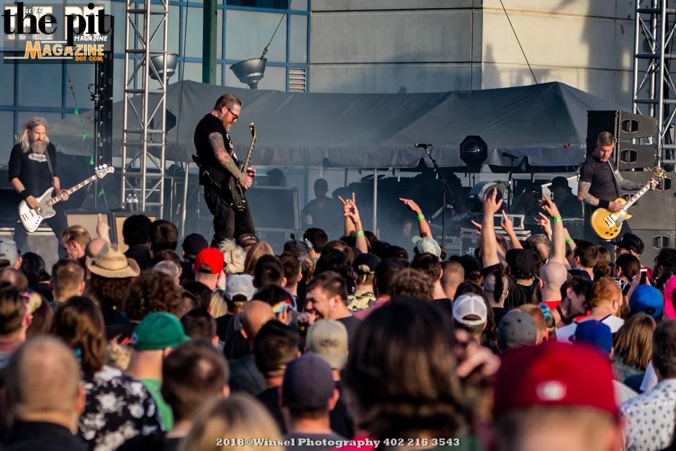 A rock band, Mastodon, performs on an outdoor stage in front of an enthusiastic crowd with raised hands, capturing a lively concert atmosphere.