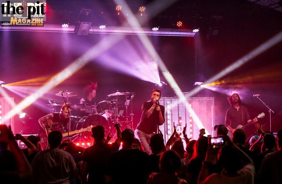 A rock band, including the renowned group Hinder, performs live on stage, illuminated by vibrant purple and white stage lights, with an engaged audience in the foreground capturing the moment on their phones.