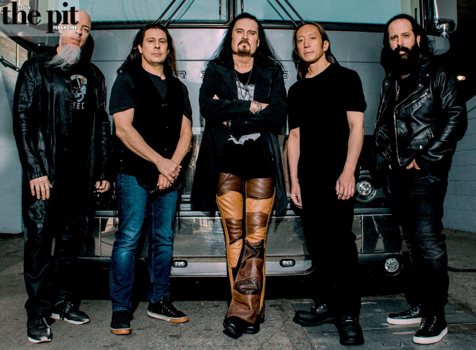 Five members of Dream Theater, standing together in a gritty urban setting, dressed in black and leather outfits.