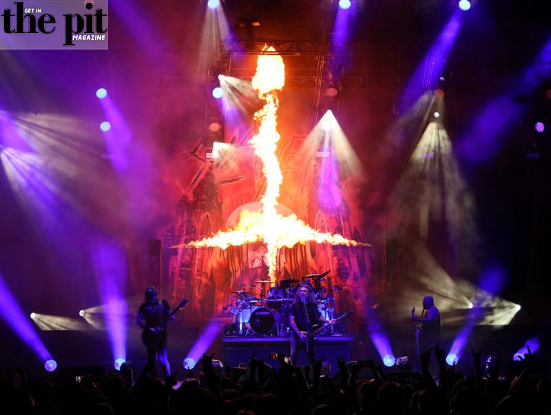 A heavy metal band performing on stage with a fiery backdrop and dramatic purple lighting, to an audience visible in the foreground.