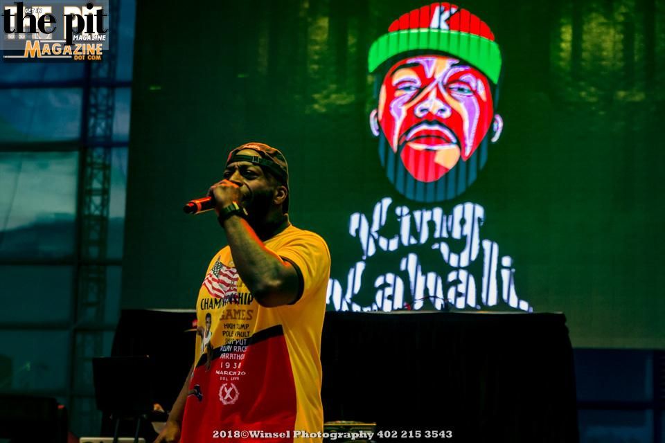A male performer in a yellow t-shirt and red cap singing on stage with a vivid green background displaying King Kahali.