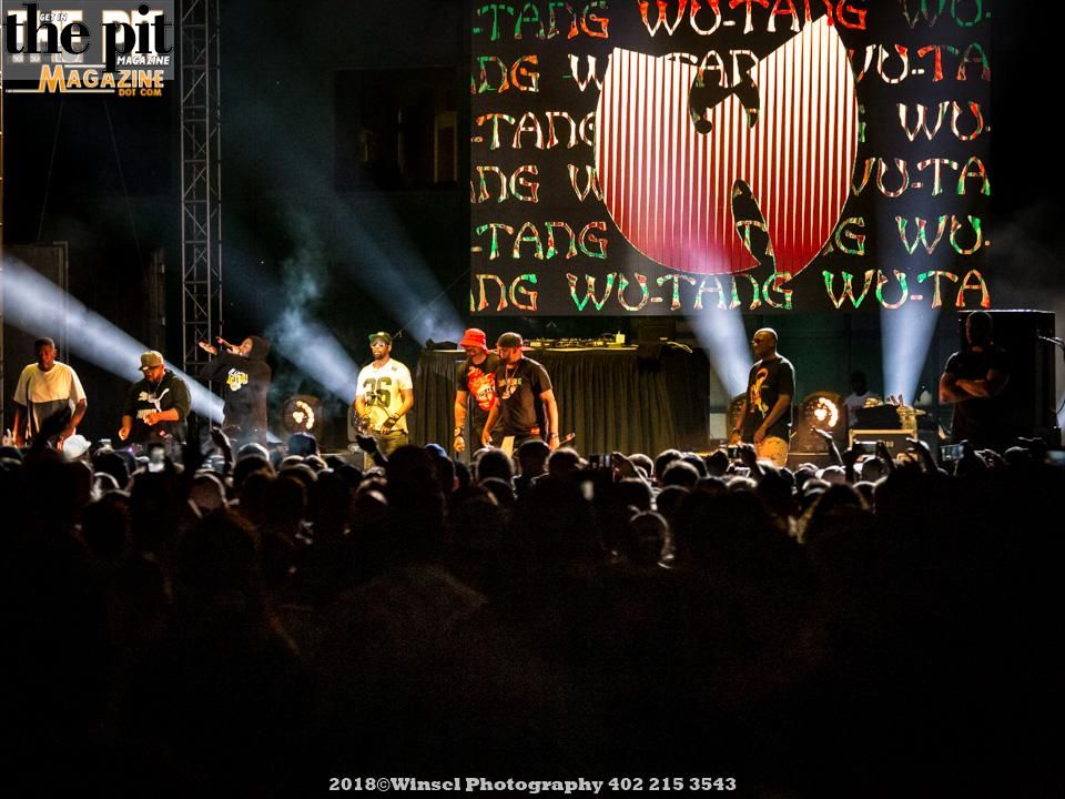 Wu Tang Clan performing on stage under a spotlight with a large Wu Tang Clan logo in the background, audience visible in the foreground.