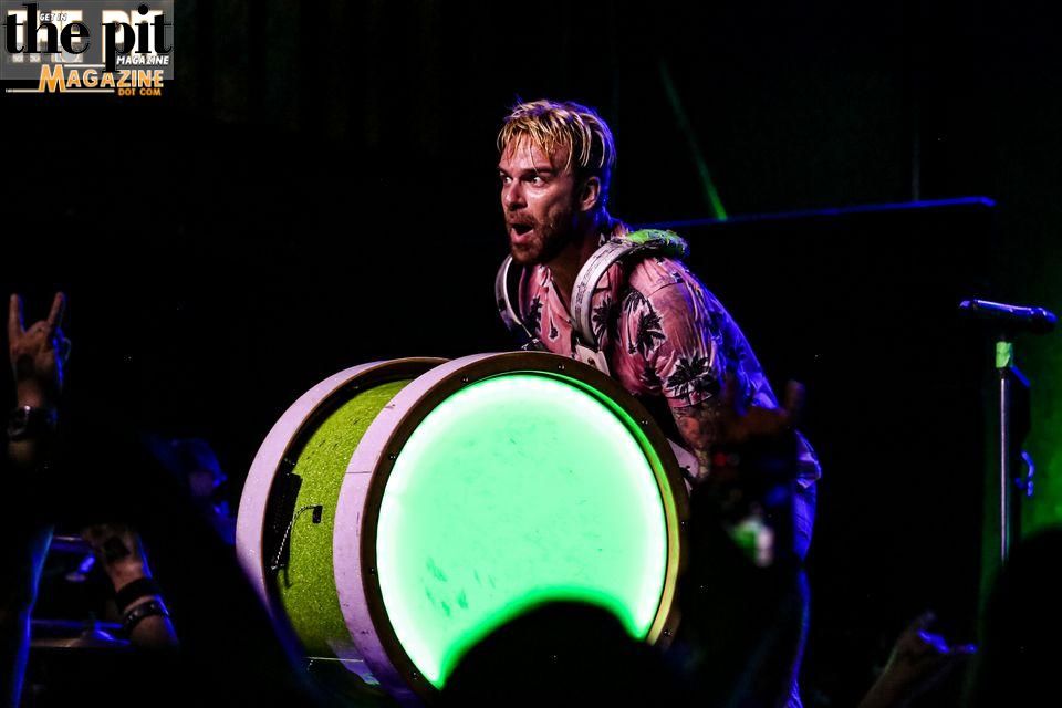 A man with spiked blonde hair intensely plays a glowing green drum onstage at a concert, with "Wilson's Pit Magazine" logo visible at the top.