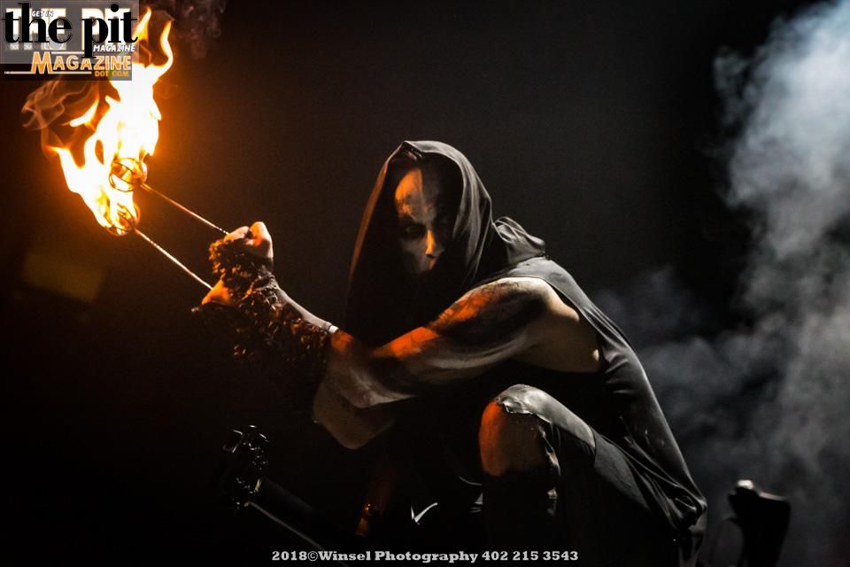 A performer in a hooded cloak breathes fire from a torch in a dimly lit setting, surrounded by behemoth clouds of smoke.