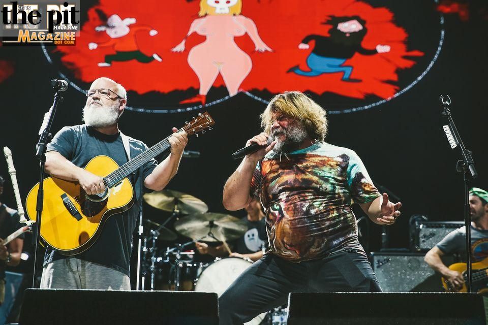 Two men from Tenacious D performing on stage, one playing an acoustic guitar and the other singing into a microphone, with a colorful animated backdrop.