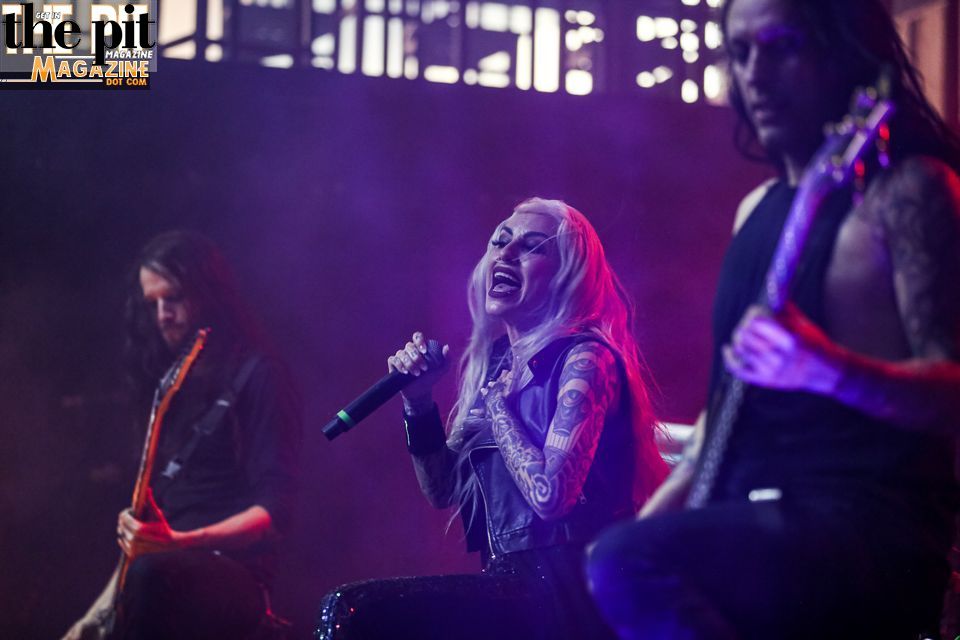Mixi Demner, a female vocalist with blonde hair and tattoos, performs on stage flanked by two male guitarists, under blue and purple stage lighting.