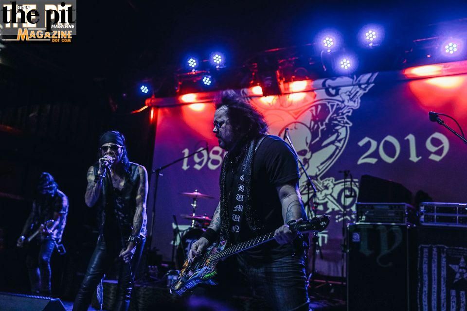 Two musicians from Bang Tango perform on stage, one singing and the other playing guitar, under blue stage lights with a banner displaying "1983-2019" in the background.