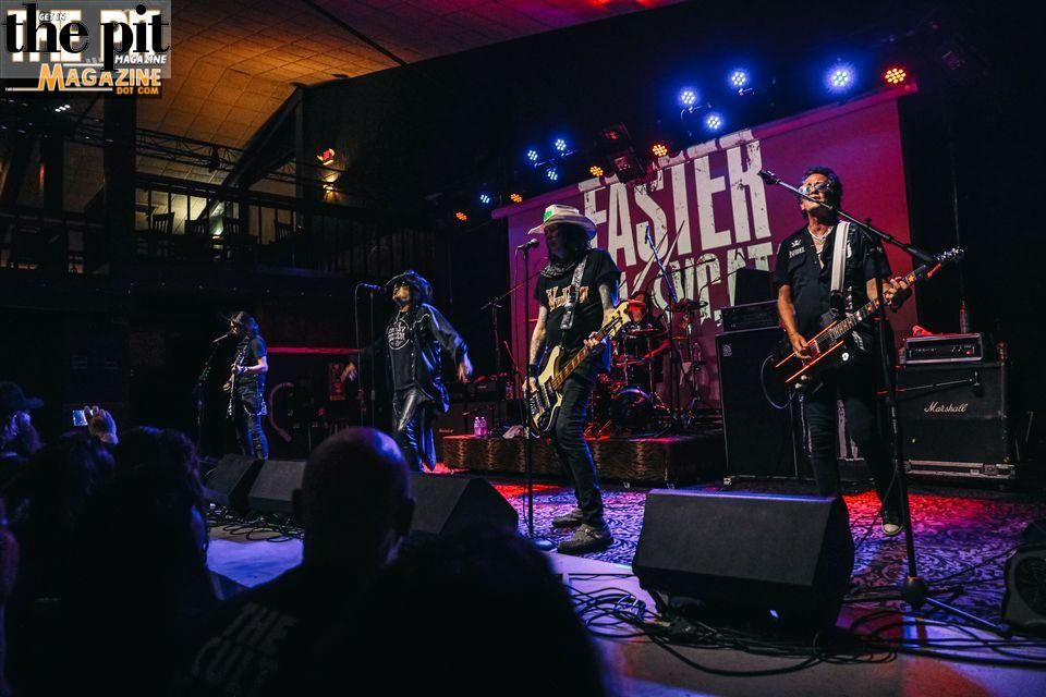 Band performing live on stage at a Faster Pussycat concert venue with blue lights reading "faster pussycat" in the background, audience in foreground.