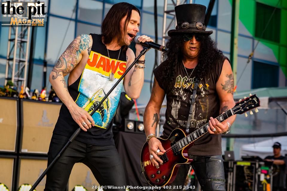 Two male rock musicians from Slash w/Myles Kennedy & The Conspirators performing on stage, one singing and one playing an electric guitar, with a crowd visible in the background.