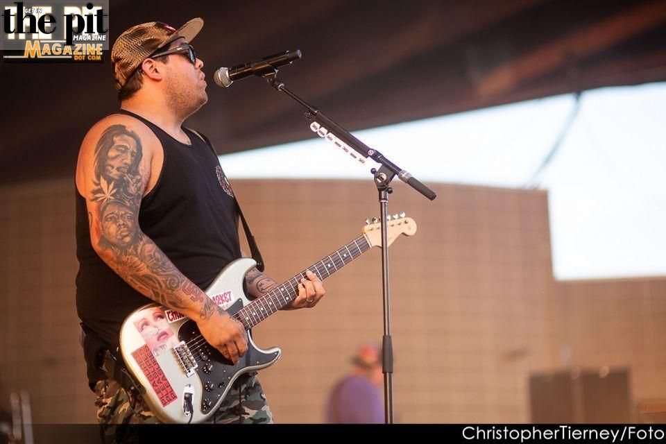 Musician with tattoos playing a white guitar on stage, wearing a black tank top and a cap, with another Sublime with Rome band member in the background.