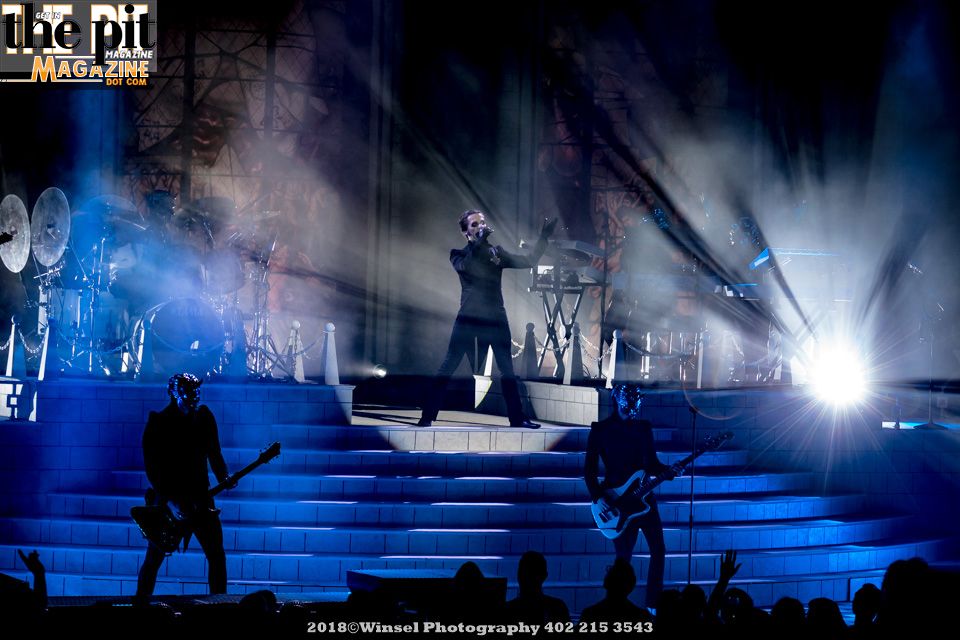 A live concert scene with a lead singer center stage and a guitarist on the left, all illuminated by dramatic blue stage lighting and set against a backdrop resembling ancient ruins haunted by ghostly figures.