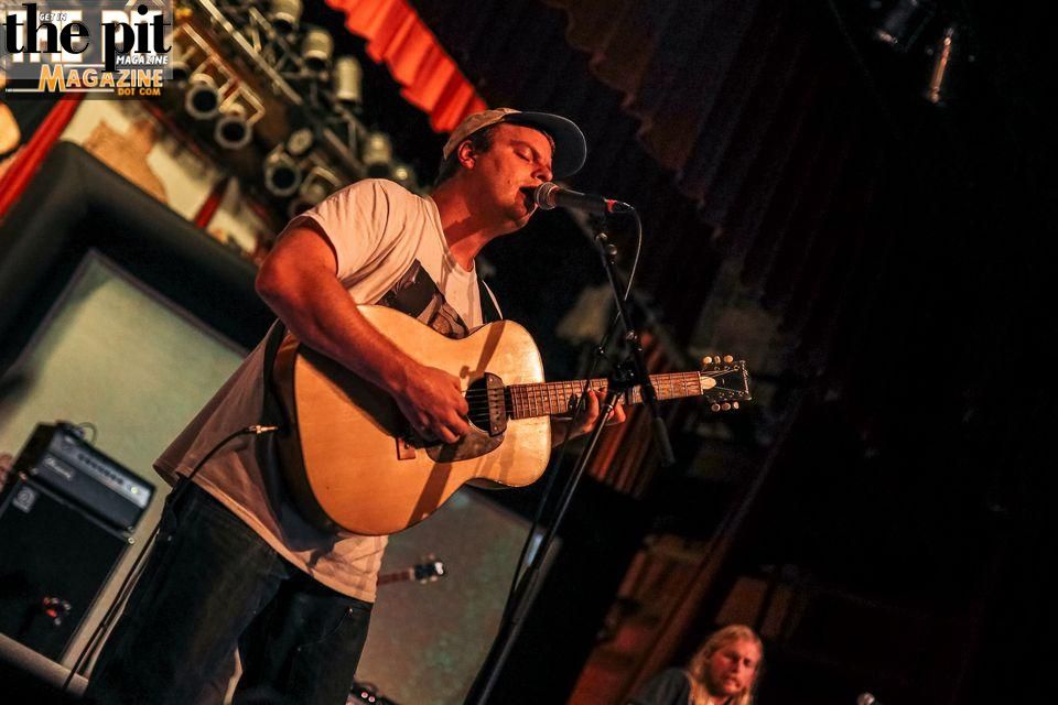 A male musician resembling Mac DeMarco in a cap plays acoustic guitar and sings into a microphone on stage, with soft lighting and another band member in the background.