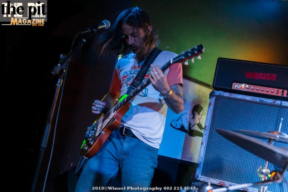 A guitarist with long hair wearing a "nola pool" t-shirt plays on stage, focused on his electric guitar, with a marshall amplifier in the background.