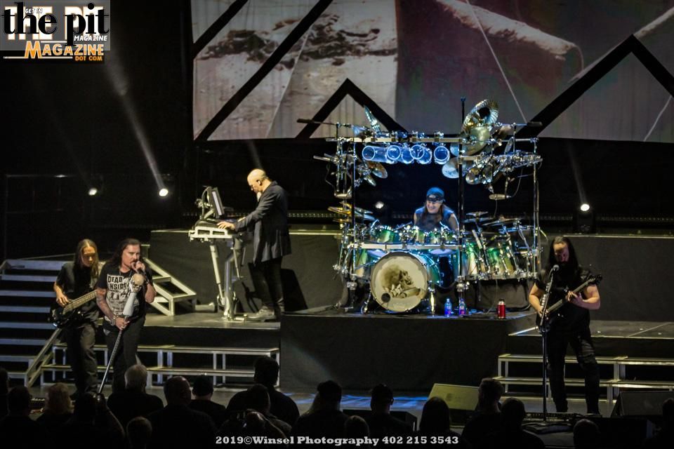 A live Dream Theater concert scene with the band performing; vocalist, keyboardist, and two guitarists on stage, with a drummer in the background under large screens.