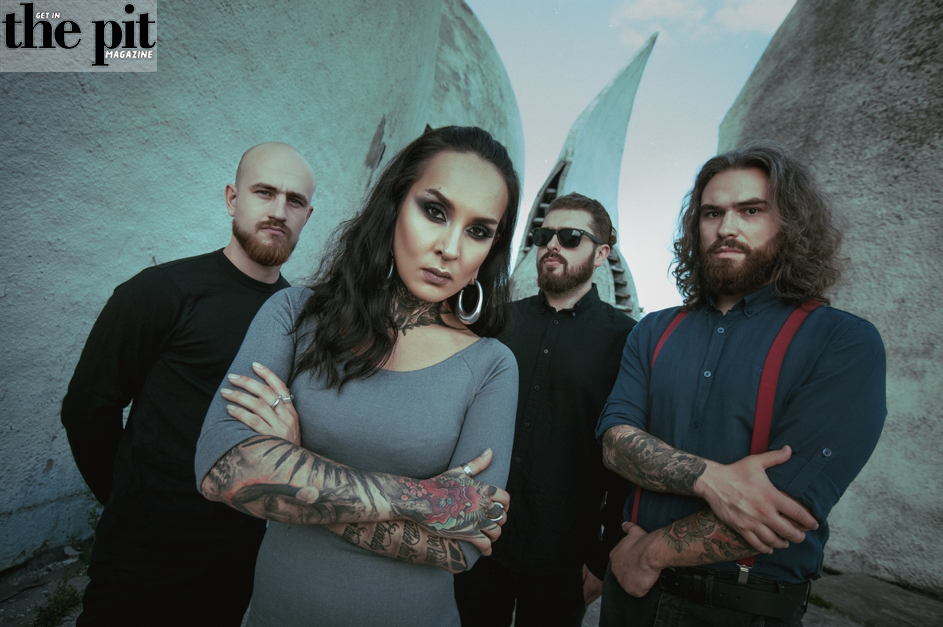 Four band members posing confidently against a sculptural backdrop, with Jinjer's lead female singer in front, featuring distinctive tattoos and a serious expression.