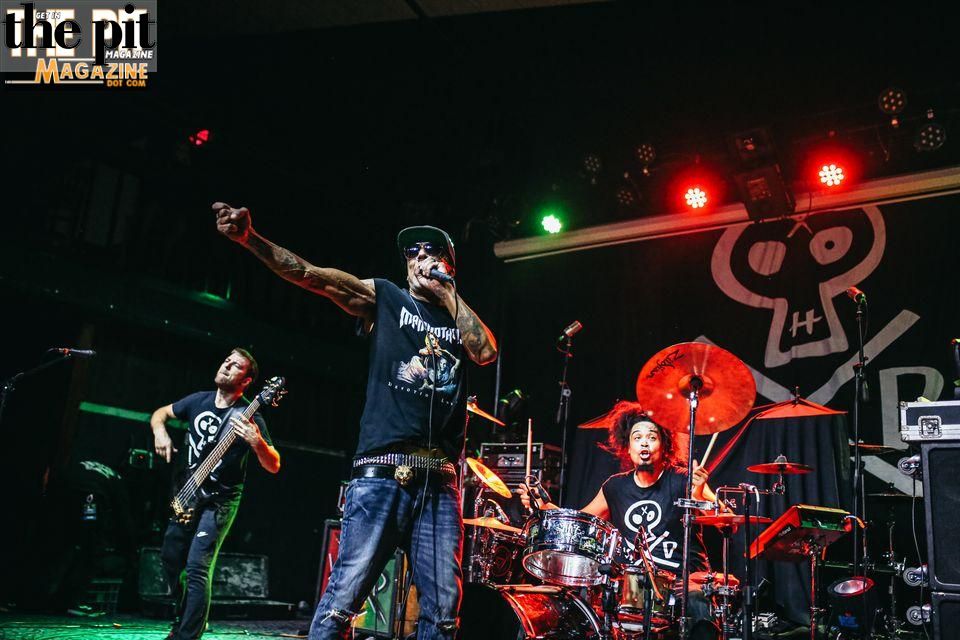 A rock band performs on stage; the lead singer gestures energetically towards the audience while the guitarist and drummer play in the background.