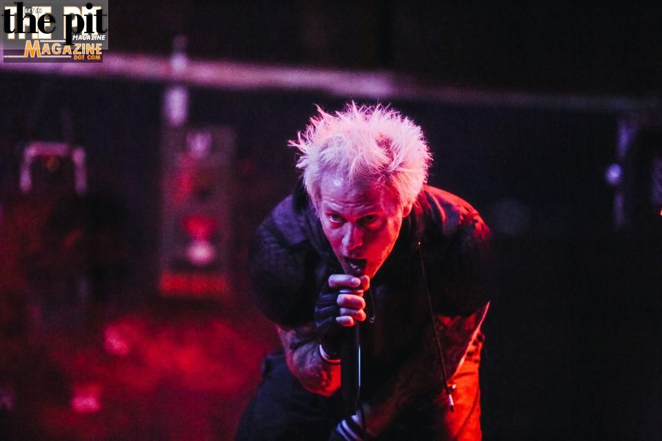 Rock musician from Powerman 5000 with spiked bleached hair performing passionately into a microphone on a dimly lit stage, highlighted by red lighting.