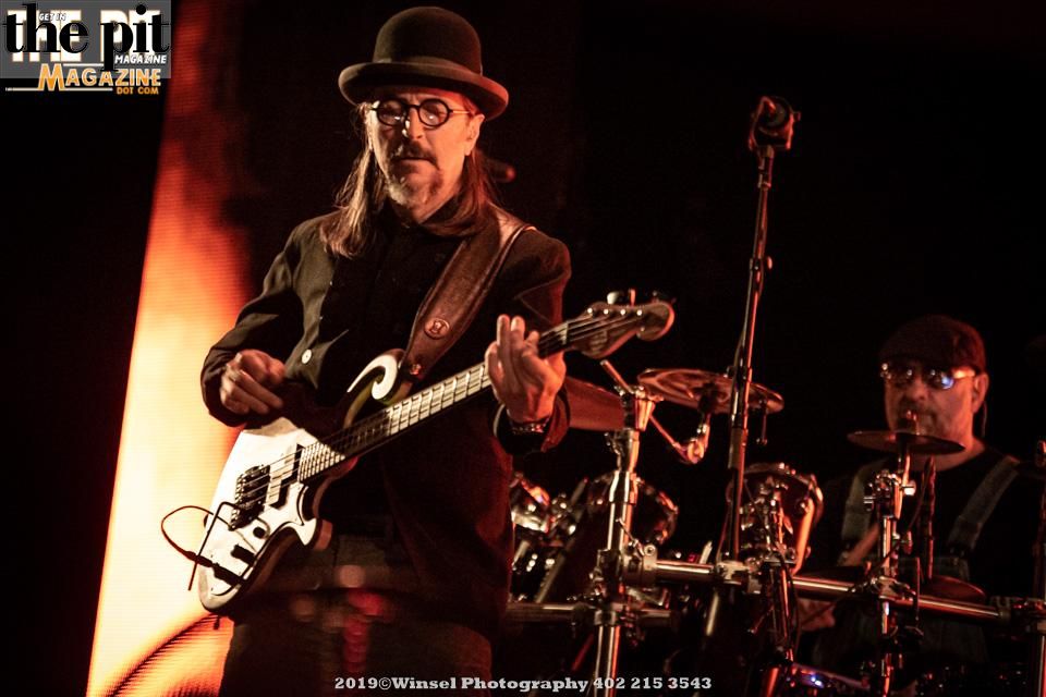 A musician in a hat and glasses plays an electric guitar on stage, illuminated by red lighting, with a drummer from Primus in the background.