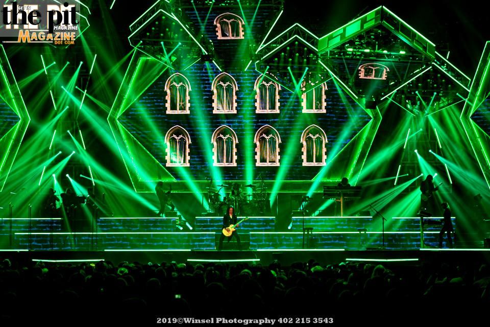 A musician from the Trans Siberian Orchestra playing guitar on stage at a concert with a cathedral-like backdrop illuminated by vivid green lights.