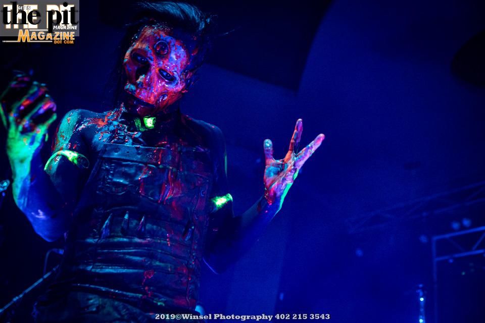Person with vibrant glow-in-the-dark paint on face and body posing under blue stage lighting at a Wednesday 13 concert.