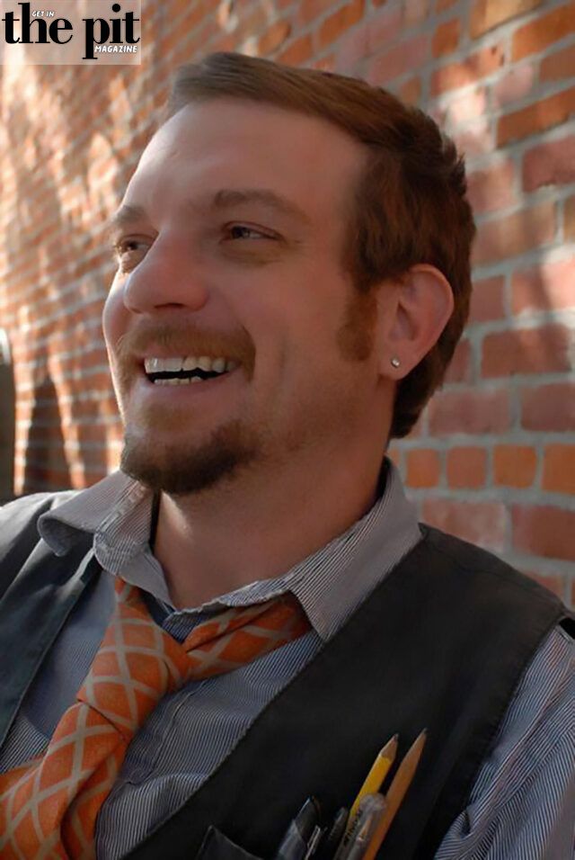 A smiling man with a light beard, wearing a vest over a dress shirt with an orange tie, stands in front of a brick wall. Mark Hennessy has a pencil tucked behind his ear.