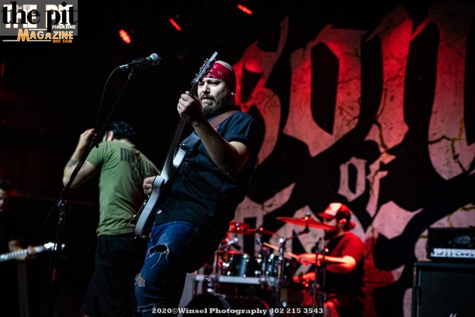 A band performing on stage, with the lead singer wearing a red bandana and holding a microphone stand, and a guitarist in the background. The backdrop features a stylized skull graphic representing Sons of Texas