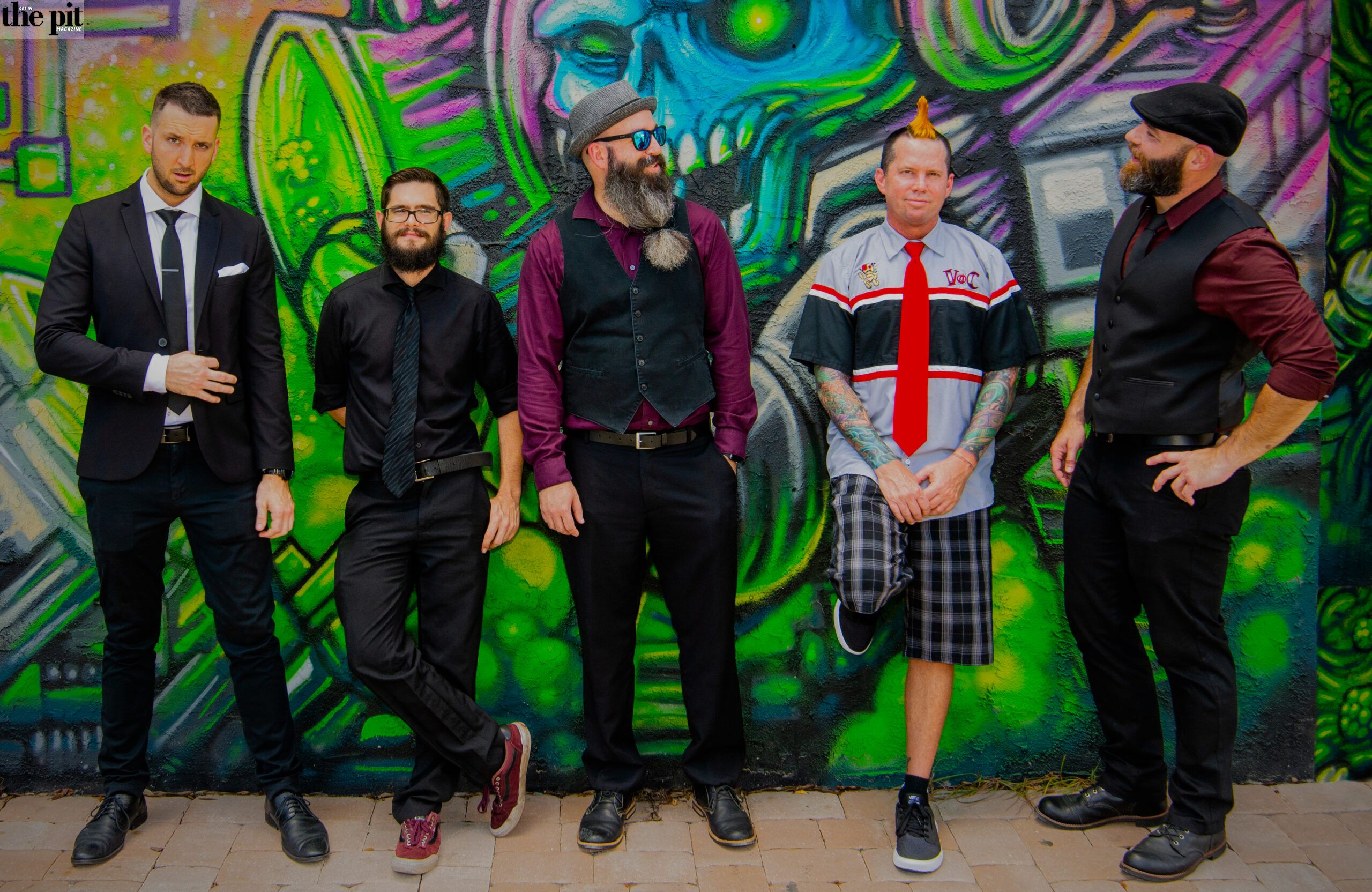 Five men in various outfits, victims of circumstance, pose in front of a colorful graffiti wall, each displaying a unique style from formal to casual.